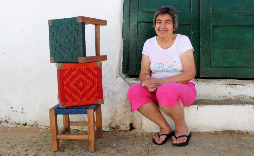Foto de María Patrocinio Pimiento, sentada en el vestíbulo de una puerta acompañada por tres artesanías tejidas en su taller AreAgua